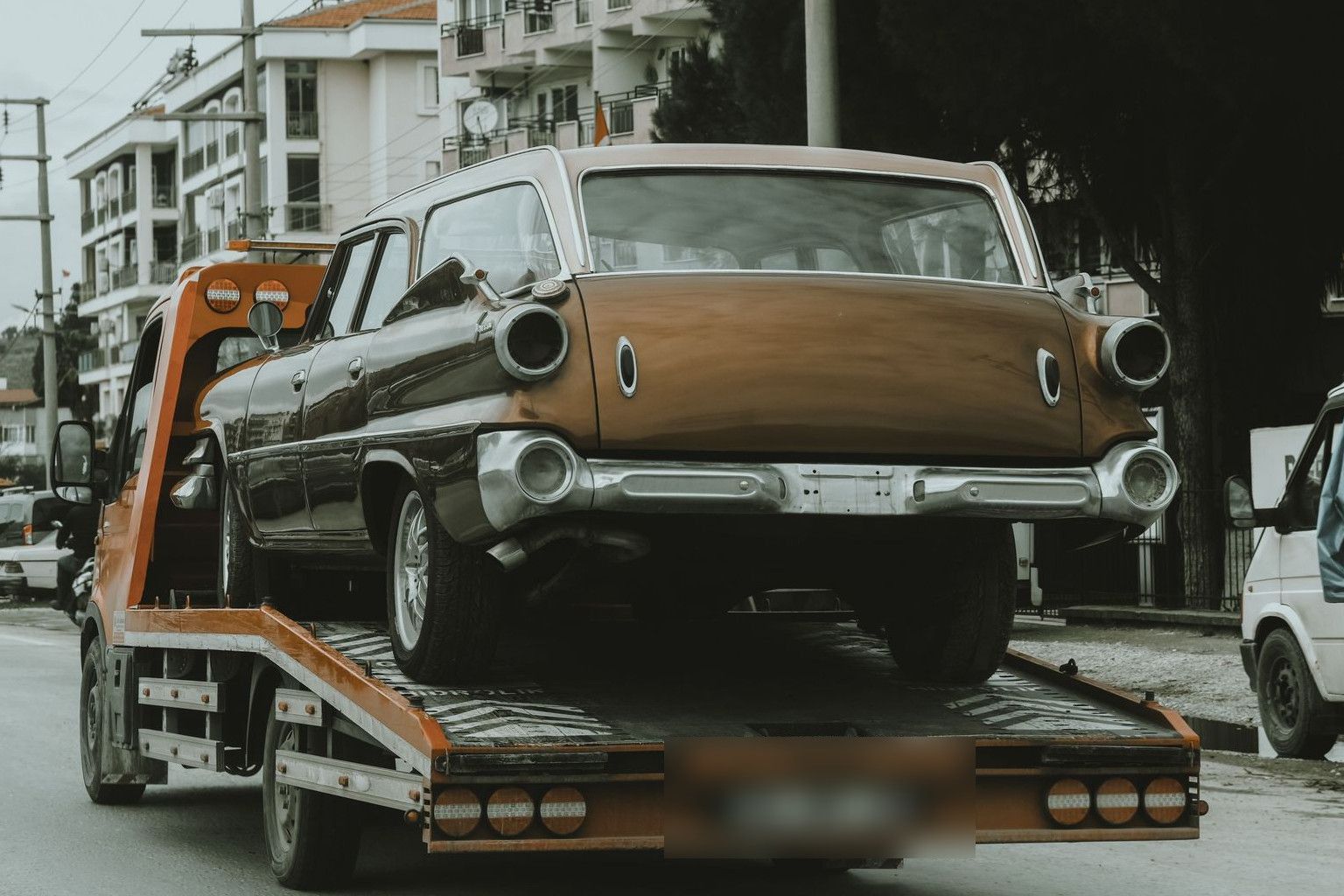 A vintage car loaded on a tow truck in a city street, surrounded by modern buildings.
